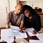 Casal sentado à mesa revisa documentos financeiros e organiza o orçamento doméstico.